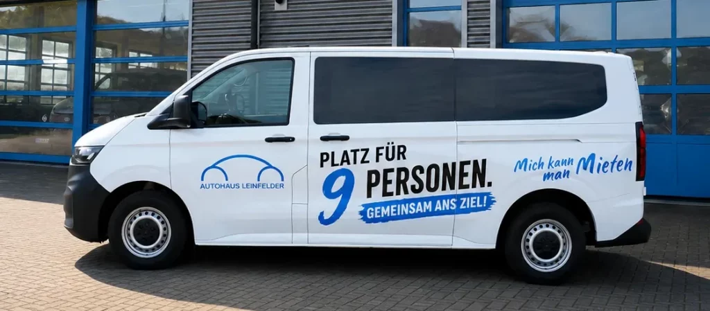 Weißer 9-Sitzer-Van mit Aufschrift "Platz für 9 Personen. Gemeinsam ans Ziel! Mich kann man mieten" vor einem Gebäude mit Schildern "Autohaus Leinfelder" und "Karosserie + Lack"